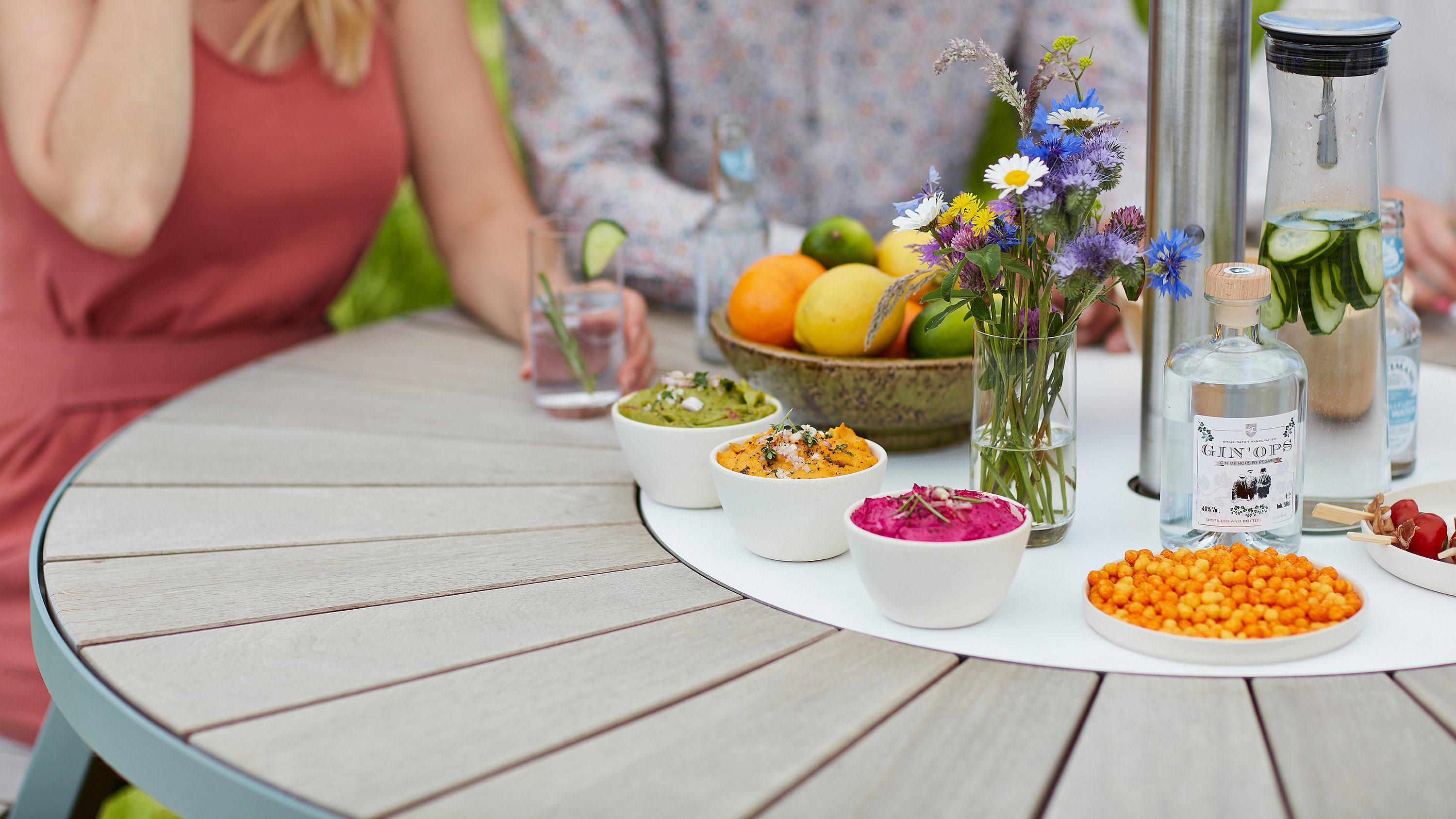 Nahaufnahme eines runden Tisches im Freien mit bunten Dips, Blumen in einer Glasvase, Zitrusfrüchten, einer Flasche Gin und einer Karaffe Gurkenwasser, um die herum Gäste sitzen.