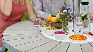Nahaufnahme eines runden Tisches im Freien mit bunten Dips, Blumen in einer Glasvase, Zitrusfrüchten, einer Flasche Gin und einer Karaffe Gurkenwasser, um die herum Gäste sitzen.