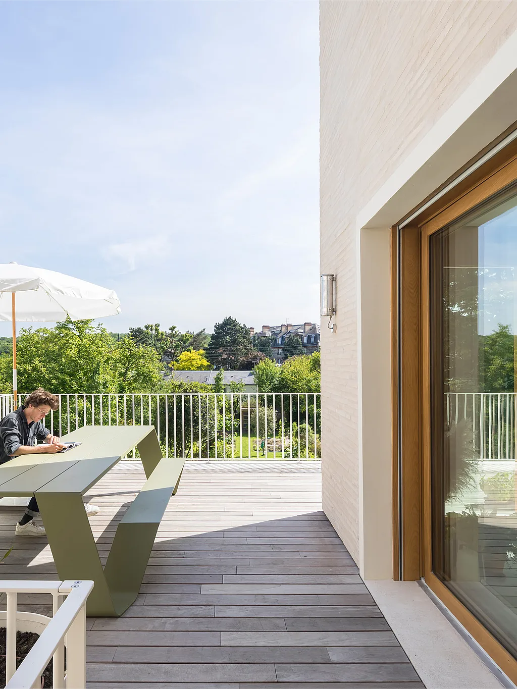 Reed green Hopper picnic table on a rooftop terrace beside a pale brick extension, where one person sits working under a white parasol with treetop views beyond the railing.