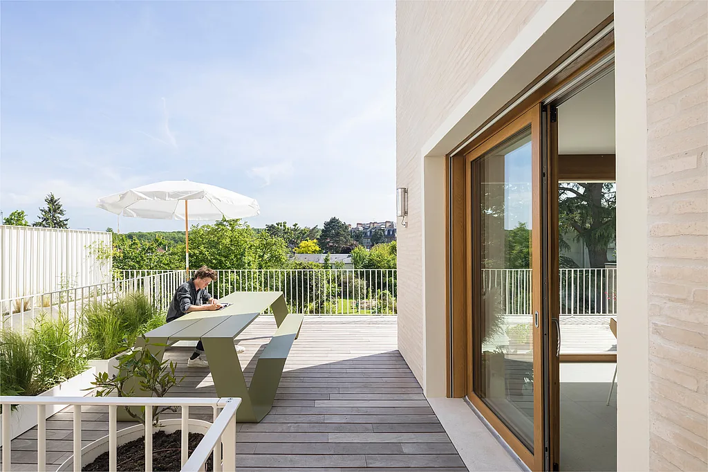 Schilfgrüner Hopper-Picknicktisch auf einer Dachterrasse neben einem Anbau aus hellem Backstein, wo eine Person unter einem weißen Sonnenschirm mit Blick auf die Baumkronen hinter dem Geländer arbeitet.