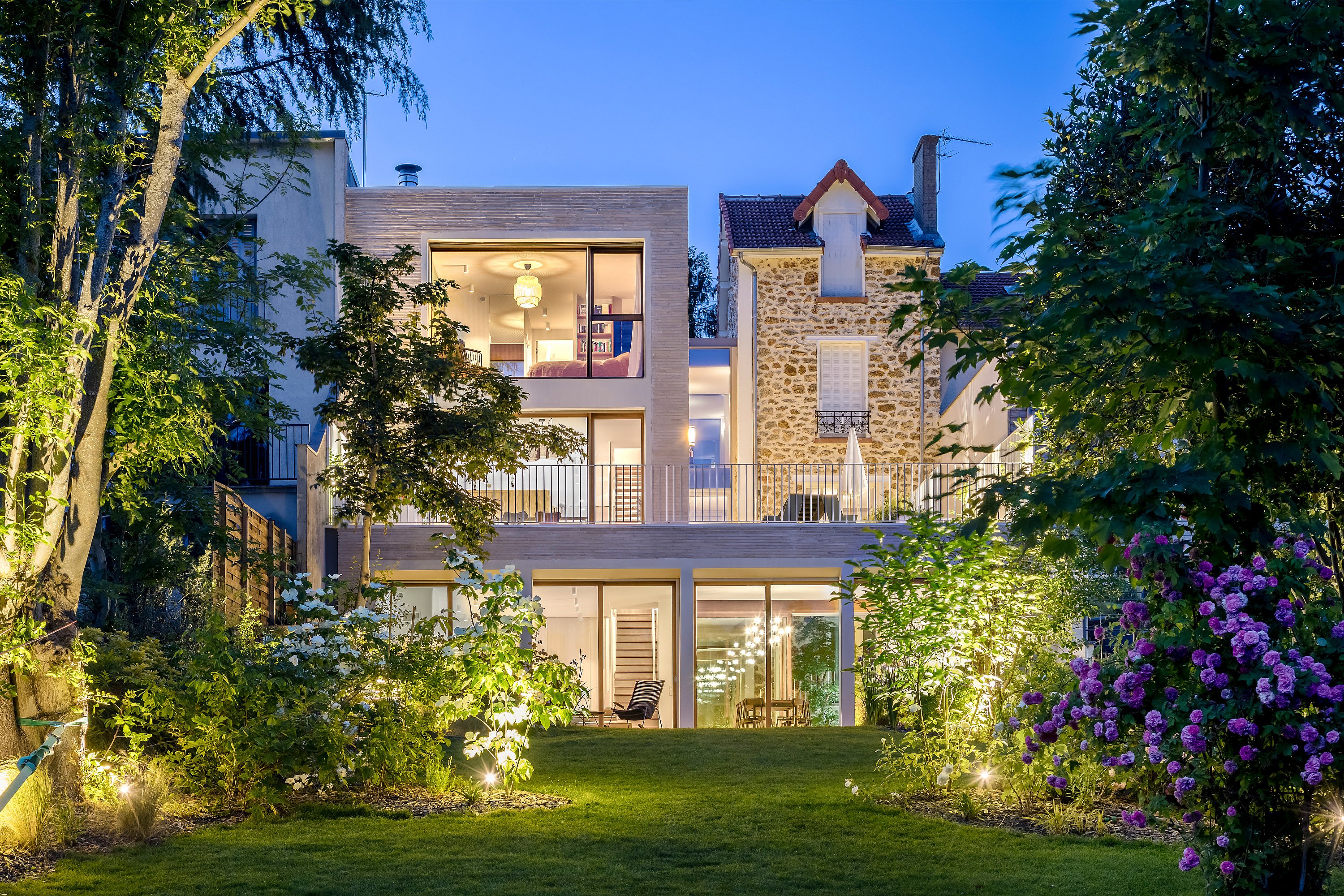 Rear view of the renovated house at dusk, showing the contrast between the old natural-stone volume and the new pale brick extension, with a rooftop terrace above a lush garden.