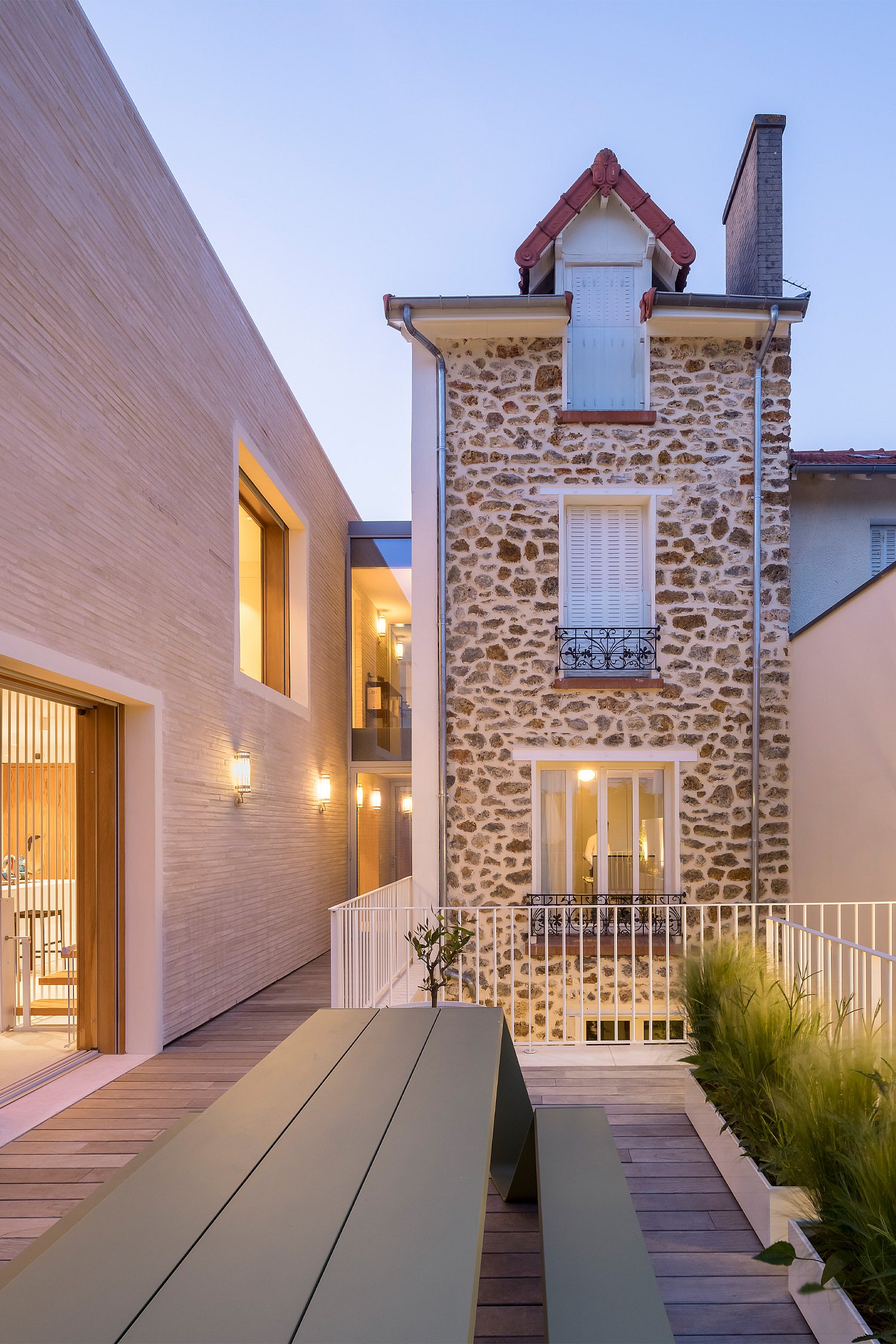 Reed green Hopper table on the rooftop terrace at twilight, set beside grasses and white railings between the new brick extension and the original stone house.