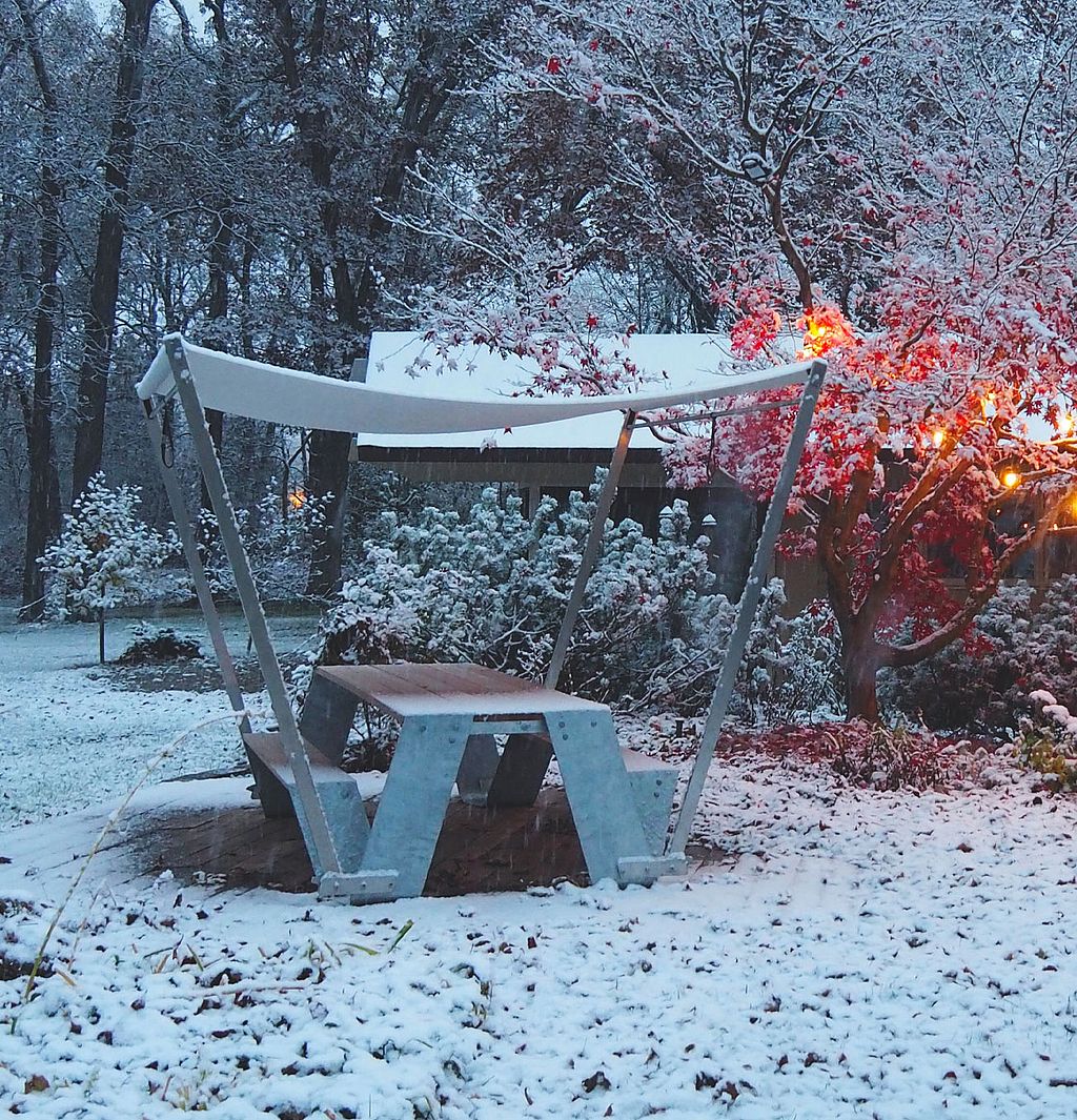 Wide view of the snowy garden in front of a low house, with a lit tree and a white Hopper table creating a cozy gathering spot in winter.