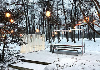 Snow-covered patio with a white illuminated Sticks divider beside a white Hopper picnic table, beneath hanging string lights in a winter forest setting.