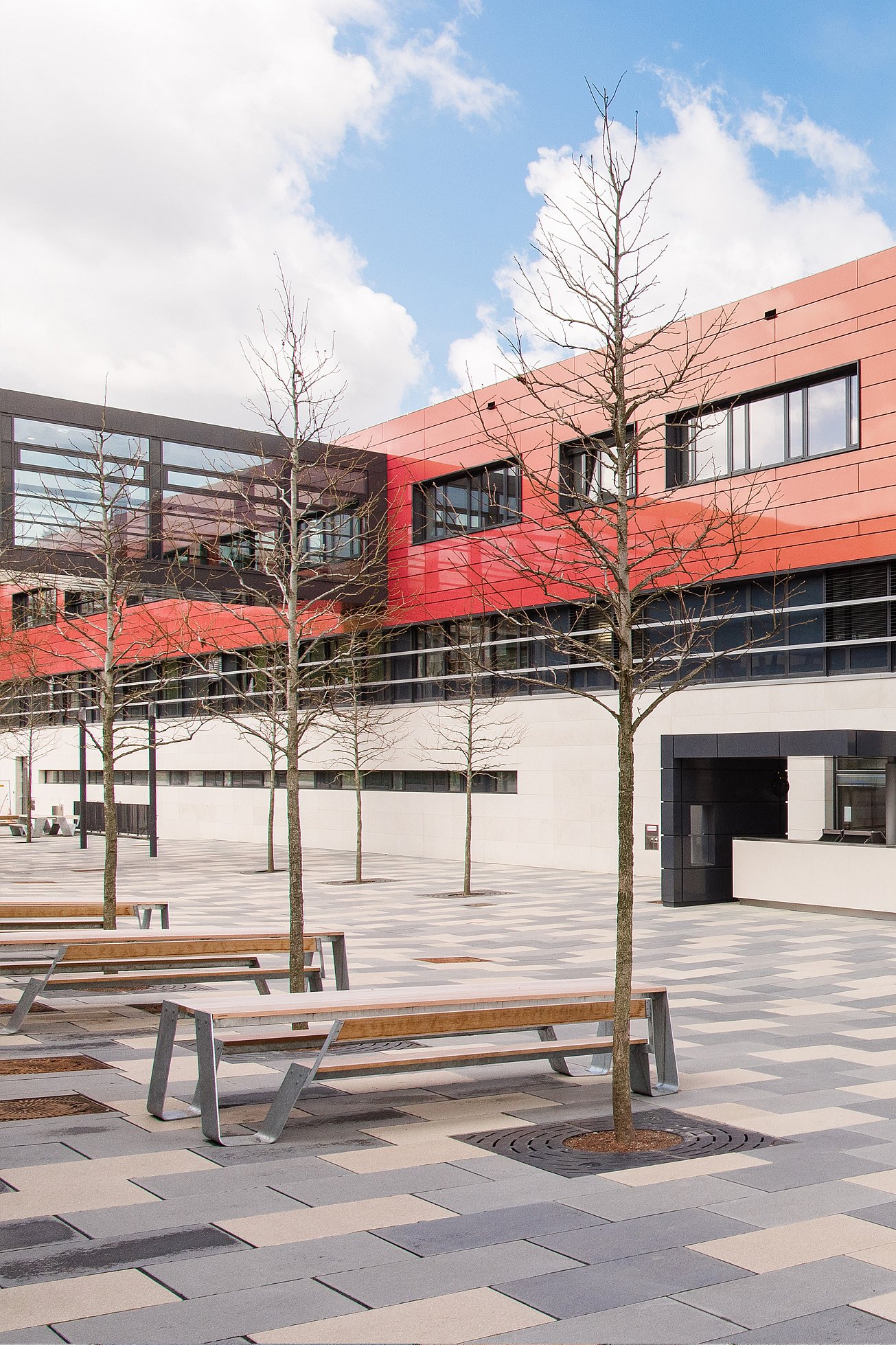 Rangée de tables de pique-nique en bois Hopper alignées sur une grande cour pavée avec de jeunes arbres, posées contre la façade rouge et orange du bâtiment de l’école sous un ciel lumineux