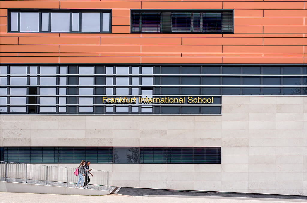 Façade de l’école internationale de Francfort avec revêtement orange, murs en pierre pâle et lettres dorées au-dessus d’une allée où deux étudiants passent.