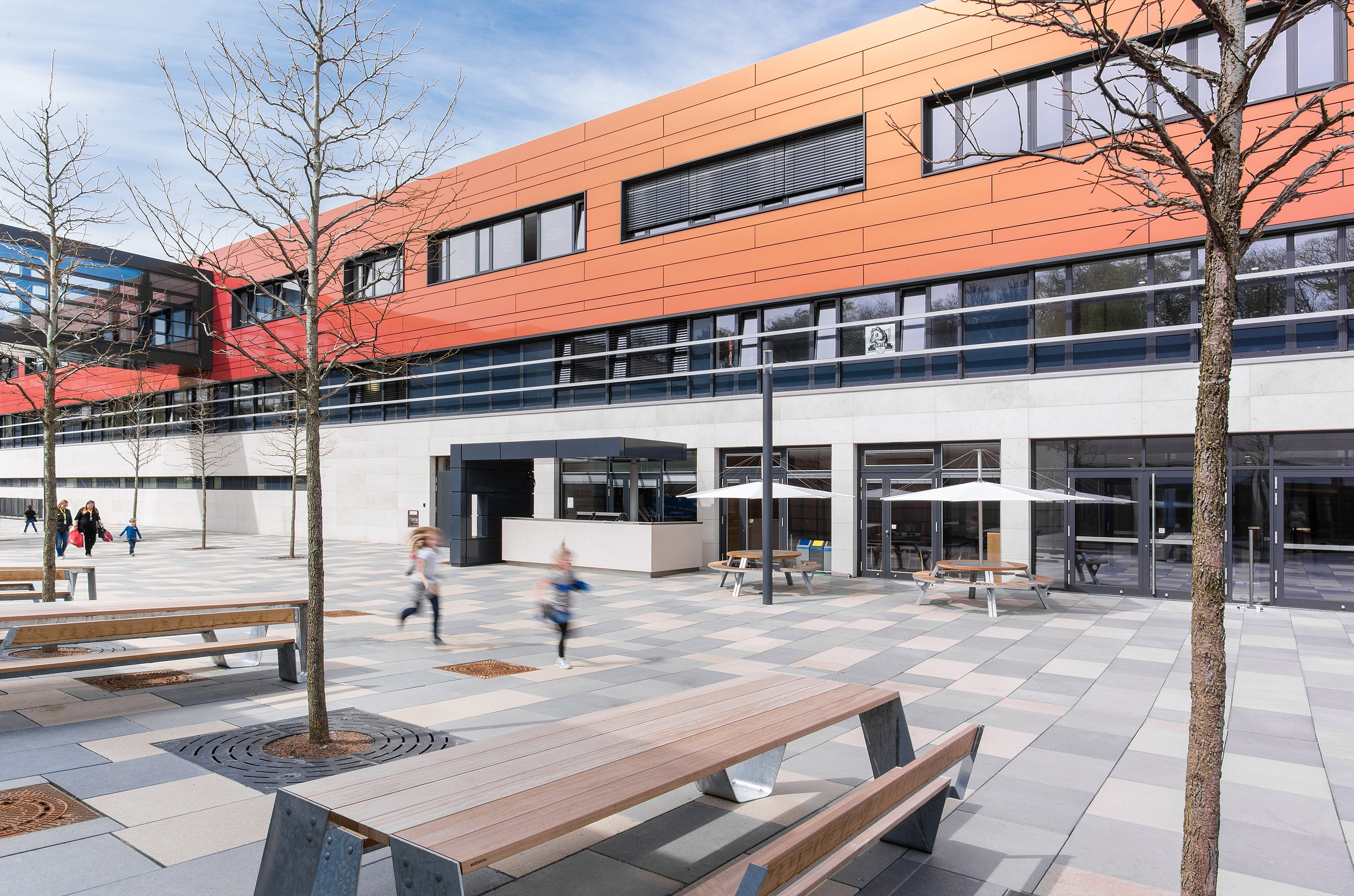 Large place d’école avec des tables de pique-nique en bois Hopper au premier plan et des tables rondes Gargantua sous des parasols blancs Inumbra près de l’entrée du bâtiment, tandis que les étudiants se déplacent à travers l’espace ouvert.