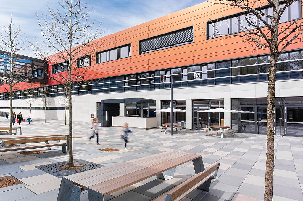 Large place d’école avec des tables de pique-nique en bois Hopper au premier plan et des tables rondes Gargantua sous des parasols blancs Inumbra près de l’entrée du bâtiment, tandis que les étudiants se déplacent à travers l’espace ouvert.