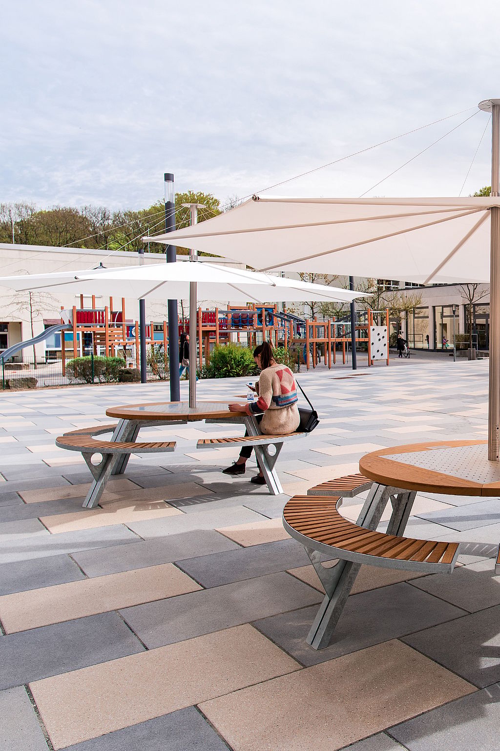 Des tables de pique-nique rondes Gargantua avec des bancs en bois à lattes sous de grands parasols blancs Inumbra dans une cour d’école ouverte à côté d’équipements de jeux colorés ; une personne est assise à travailler à une table.