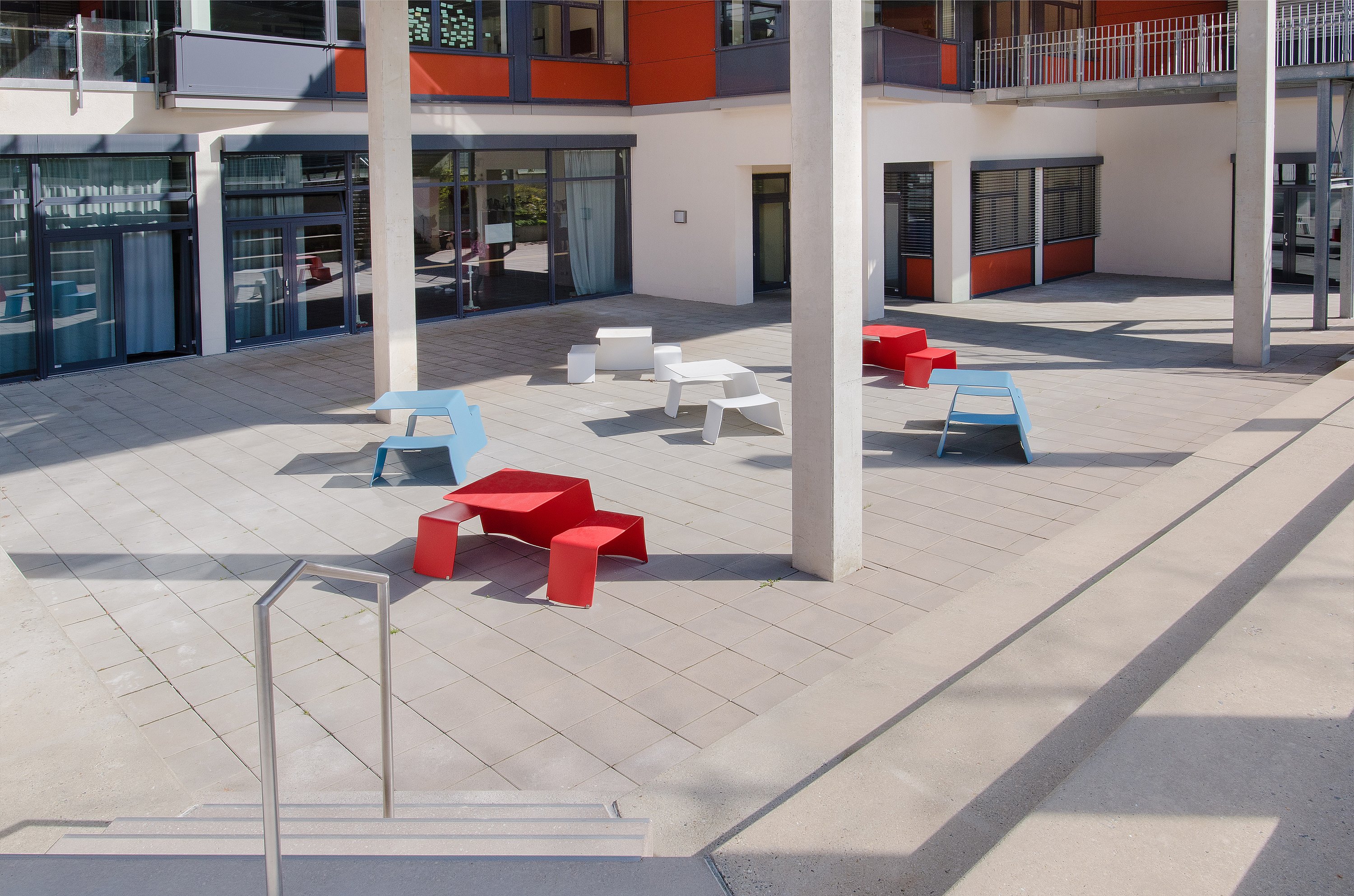 Ruime binnenplaats omlijst door een modern crème, grijs en oranje gebouw, ingericht met verspreide rode, witte en lichtblauwe Picnik tafel-en-banksets op grote bleke bestratingstegels.
