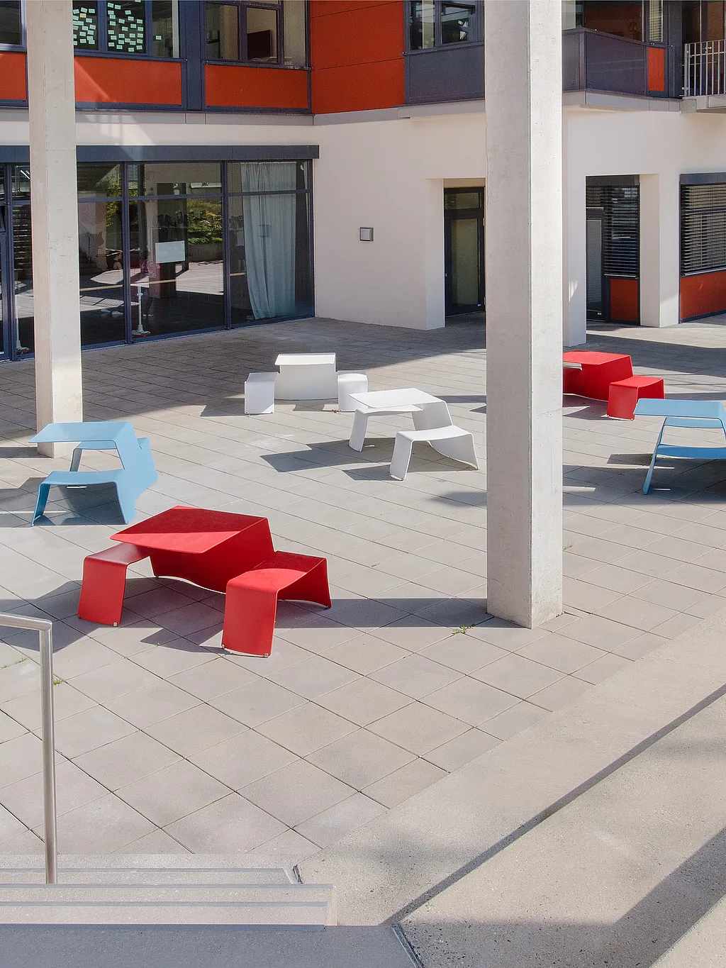 Ruime binnenplaats omlijst door een modern crème, grijs en oranje gebouw, ingericht met verspreide rode, witte en lichtblauwe Picnik tafel-en-banksets op grote bleke bestratingstegels.