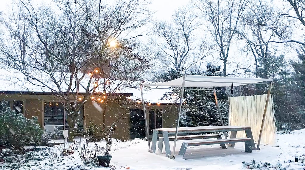 Snow-covered garden with a white Hopper picnic table beneath warm string lights, framed by trees with bright red leaves dusted in snow
