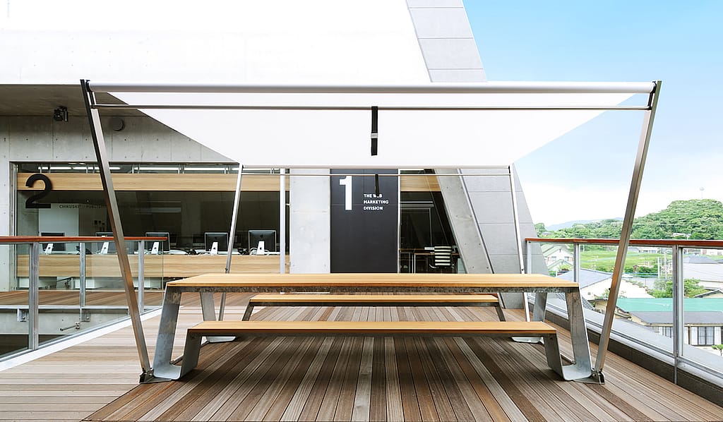 Table de pique-nique Hopper positionnée dans l’atrium aéré du niveau supérieur, offrant une vue dégagée sur la ville de Chikuho et un endroit lumineux et venteux pour s’asseoir.