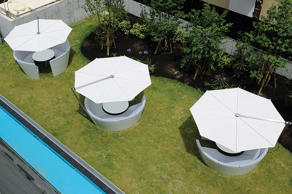 White Kosmos seating islands with large white shades arranged on a green lawn beside a black stone building, bordered by a narrow strip of bright blue water.