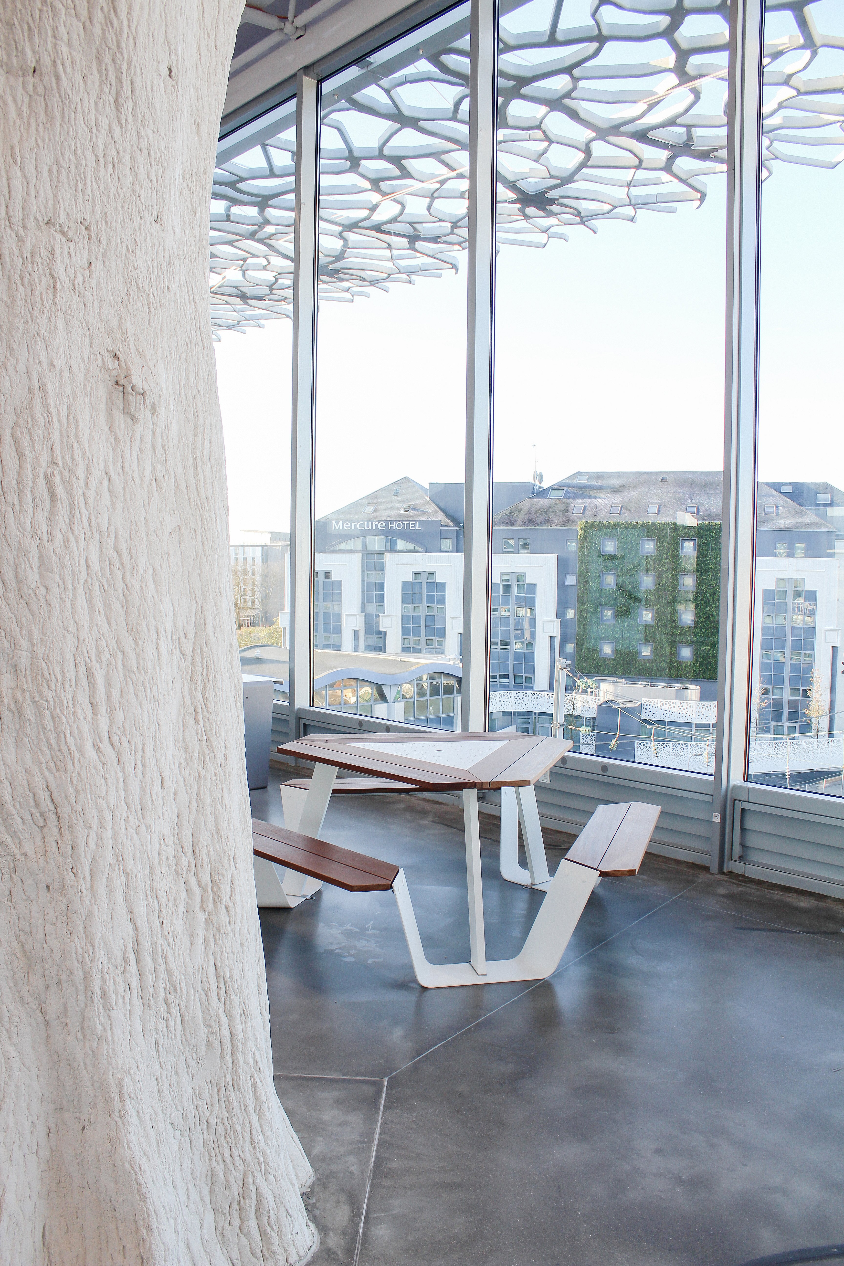 White-and-wood Anker table placed next to full-height station windows, overlooking nearby buildings beneath the patterned roof structure outside.