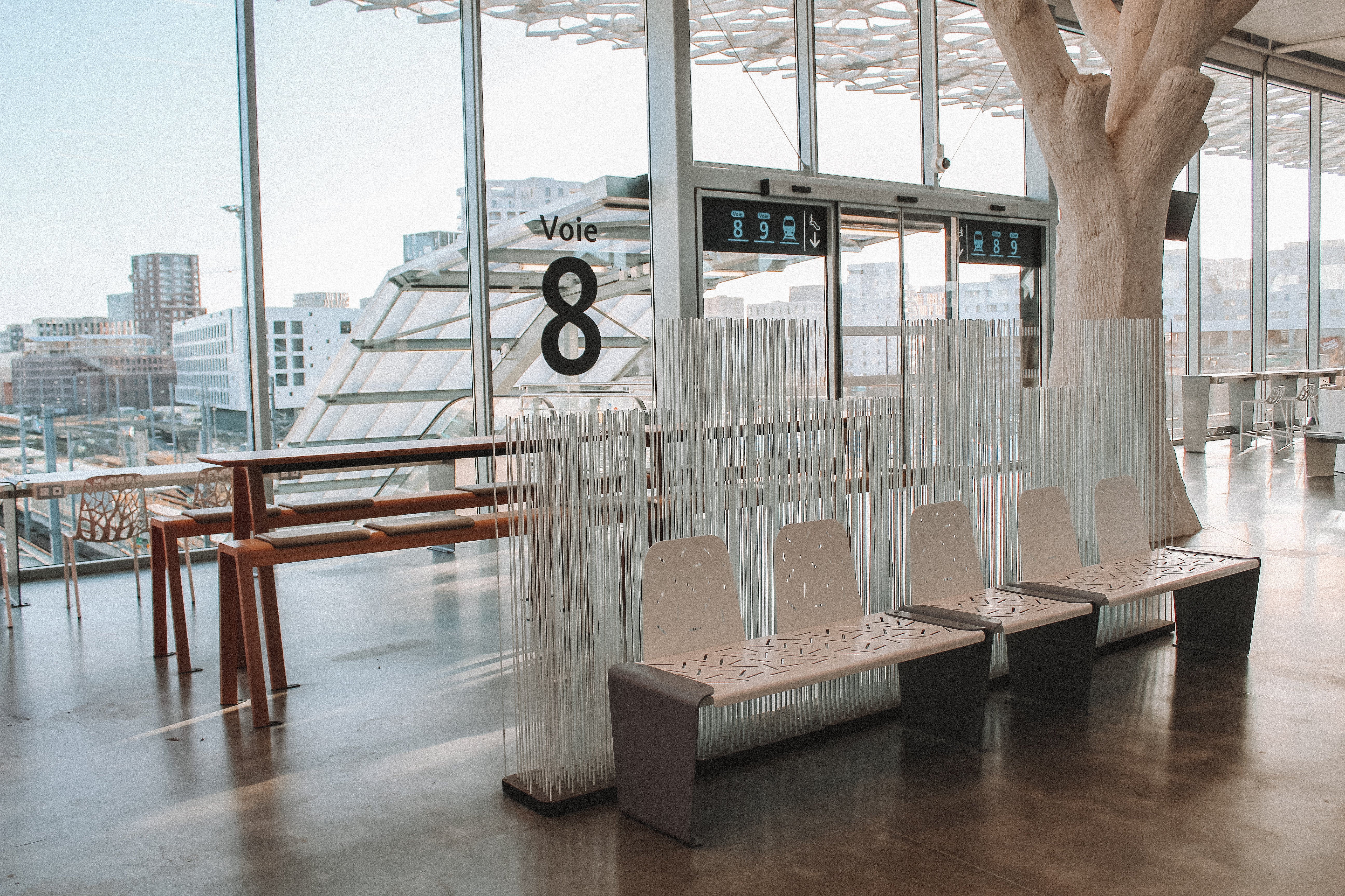 Red Anker picnic table beside a glass wall marked ‘Voie 8,’ partly screened by tall white Sticks dividers in the airy station hall.