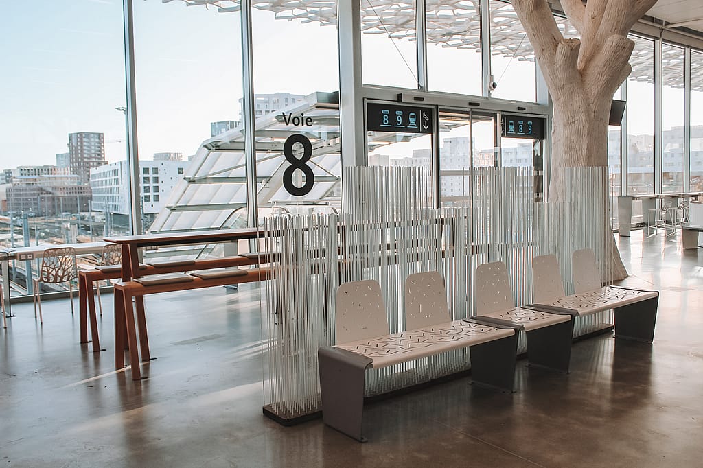 Red Anker picnic table beside a glass wall marked ‘Voie 8,’ partly screened by tall white Sticks dividers in the airy station hall.