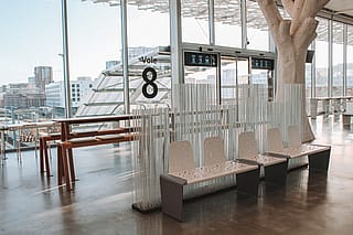 Red Anker picnic table beside a glass wall marked ‘Voie 8,’ partly screened by tall white Sticks dividers in the airy station hall.