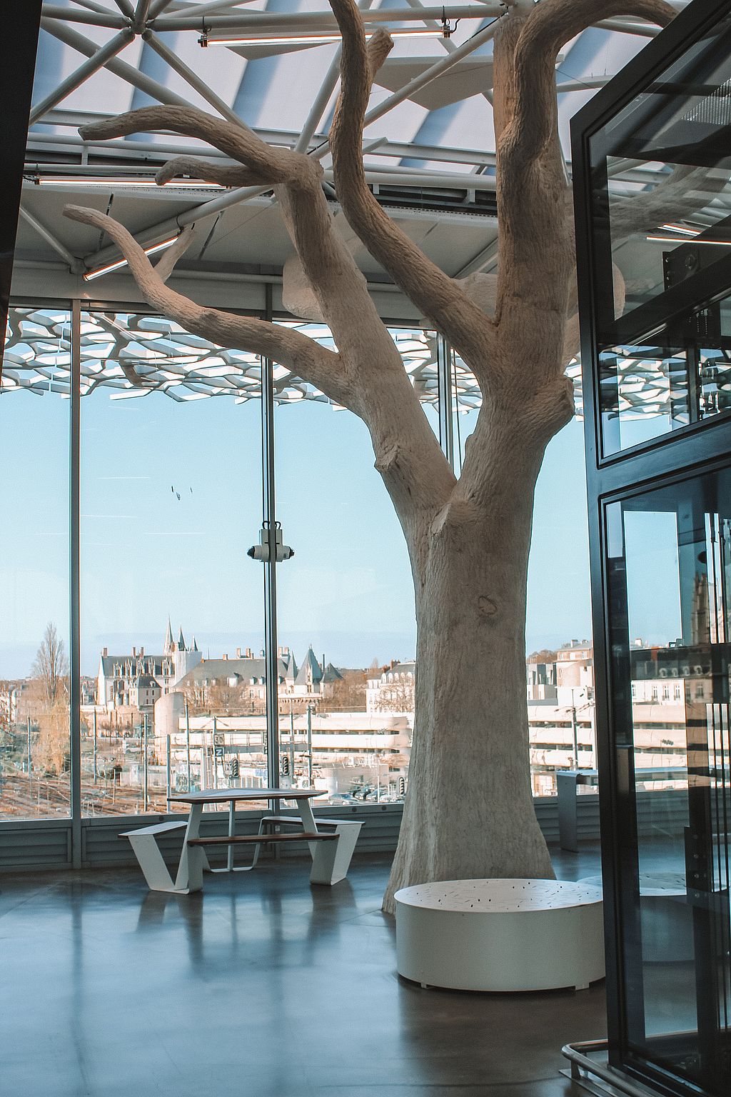 Large white concrete column shaped like a bare tree rising through the station concourse, with an Anker table and city view in the background