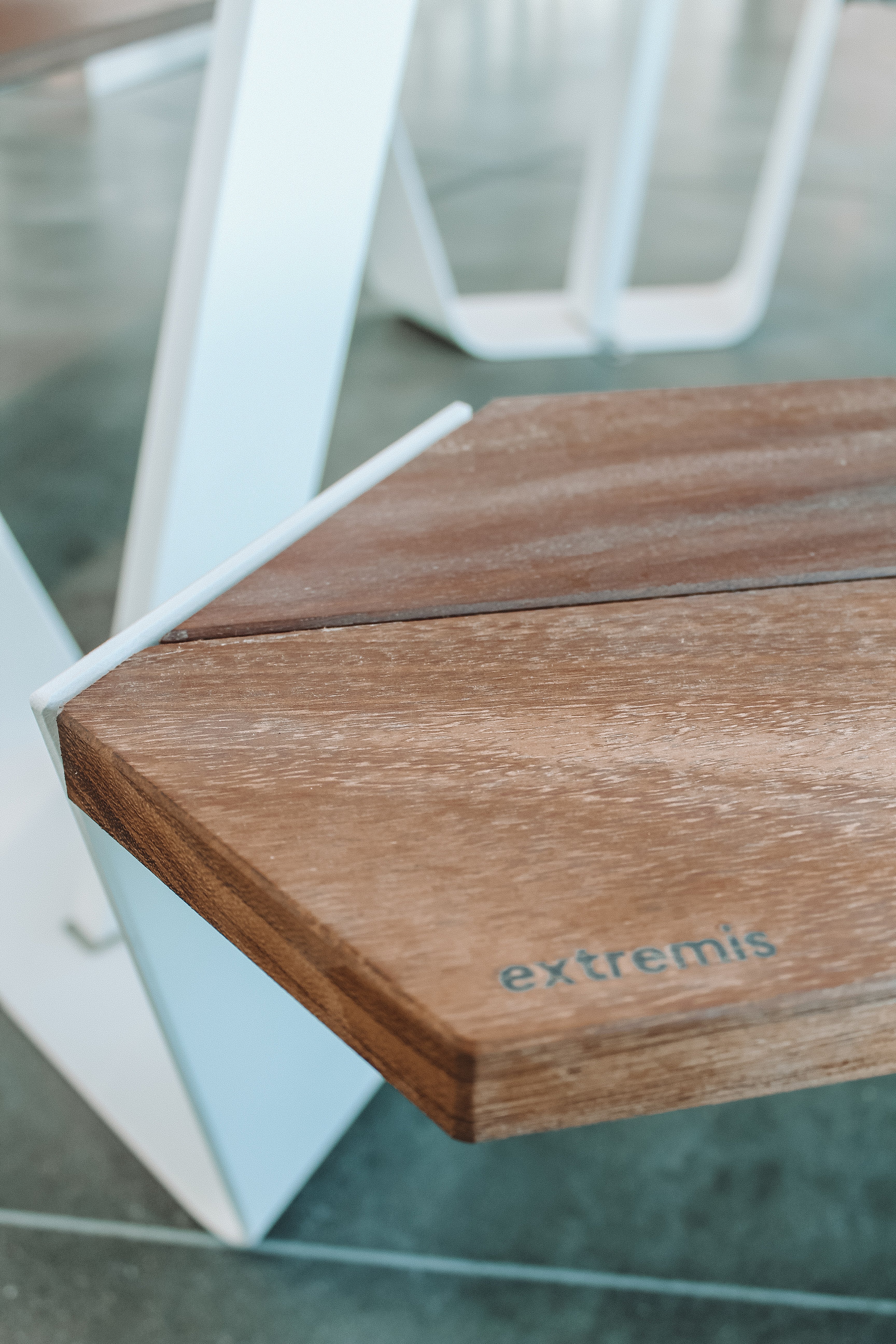 Close-up of the corner of a wooden Anker tabletop with the Extremis logo, supported by a white metal frame on the polished station floor.
