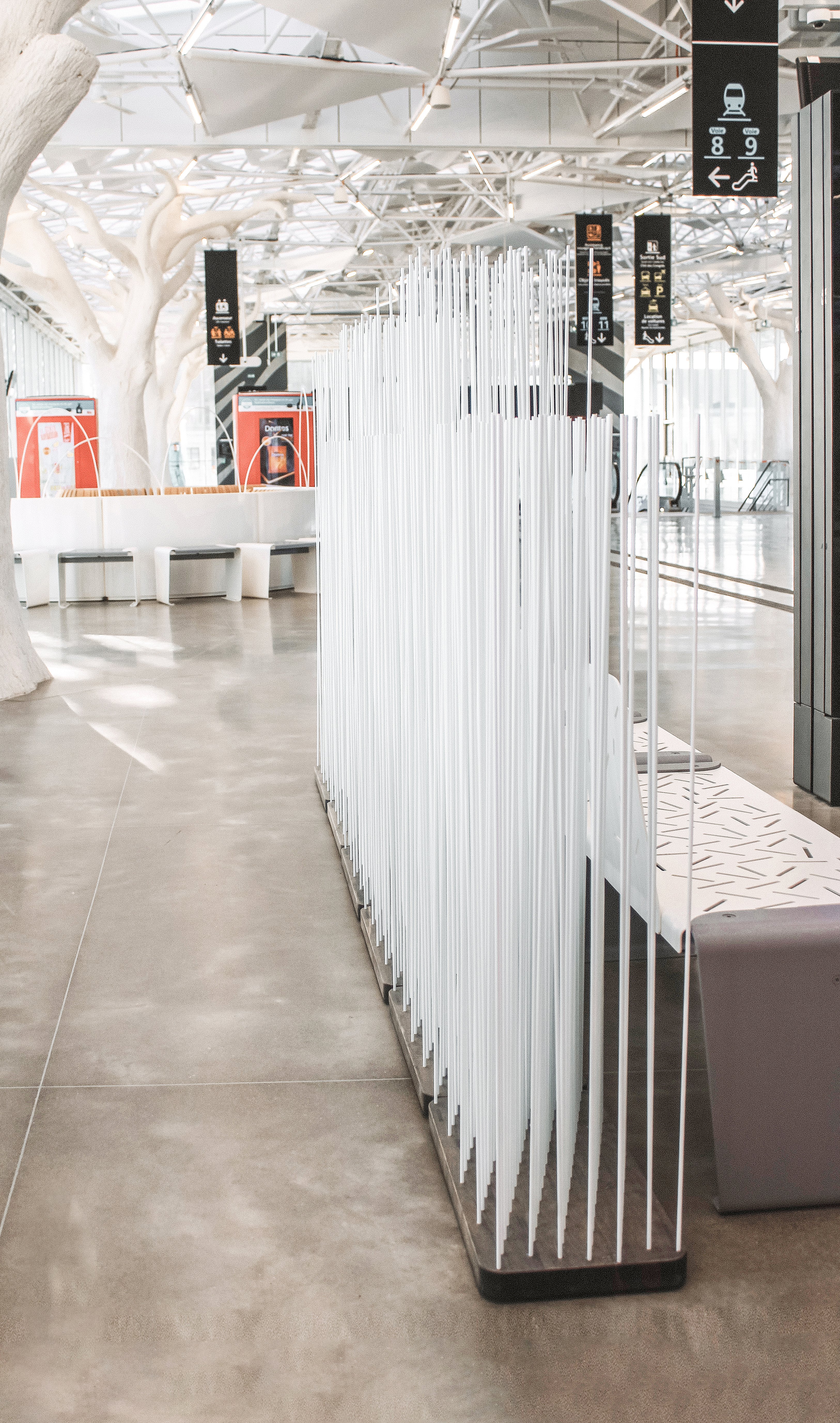 Tall white Sticks room divider standing on a dark base in the station interior, creating a light visual screen along the circulation route.