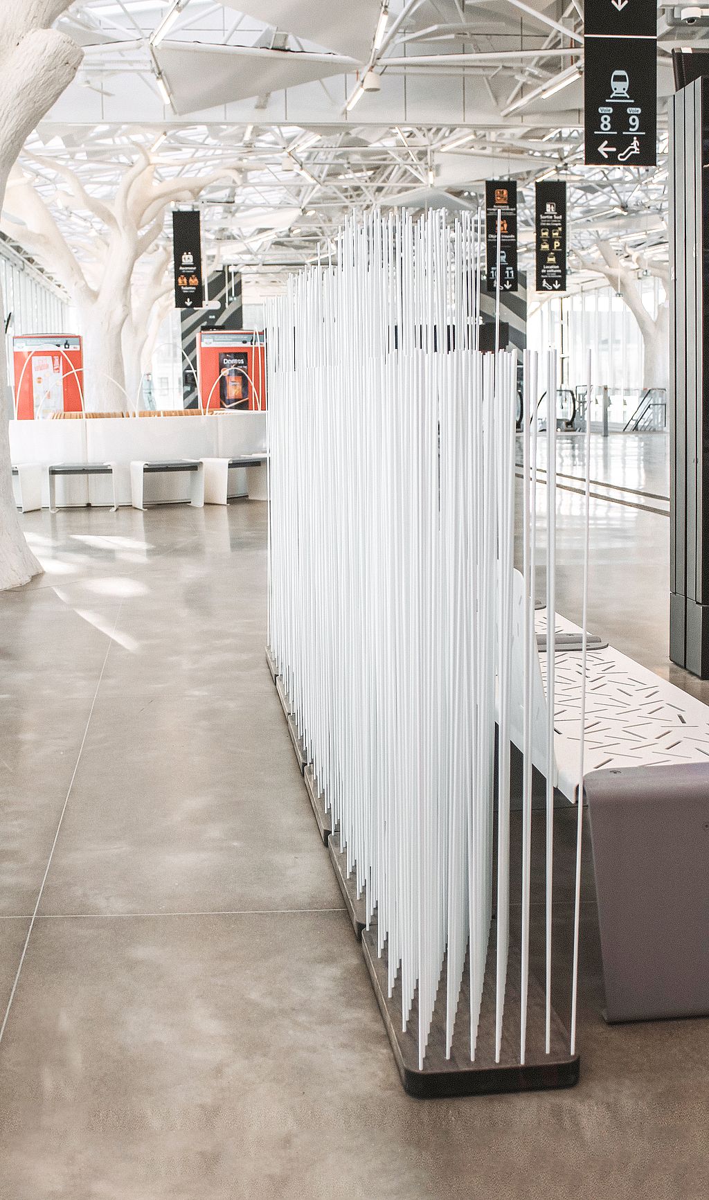 Tall white Sticks room divider standing on a dark base in the station interior, creating a light visual screen along the circulation route.