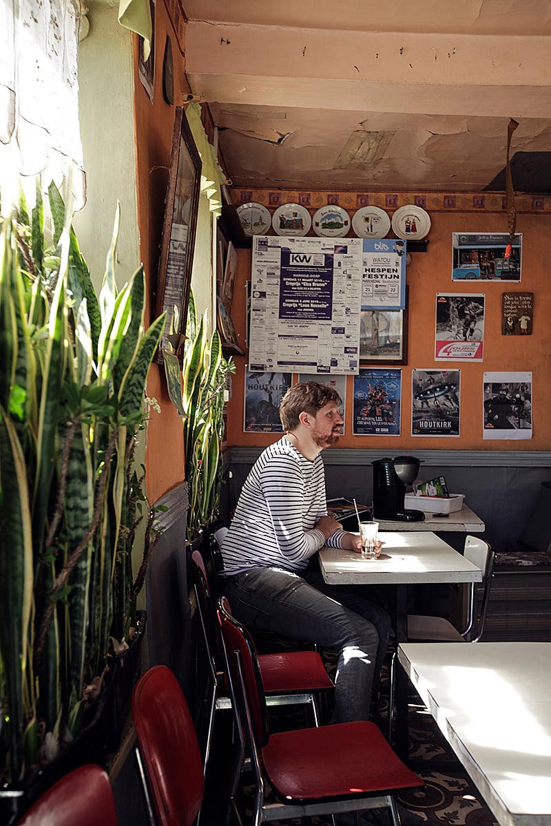 Man seated alone inside the nostalgic pub, surrounded by tall sansevierias, red chairs, vintage posters, and warm afternoon light.