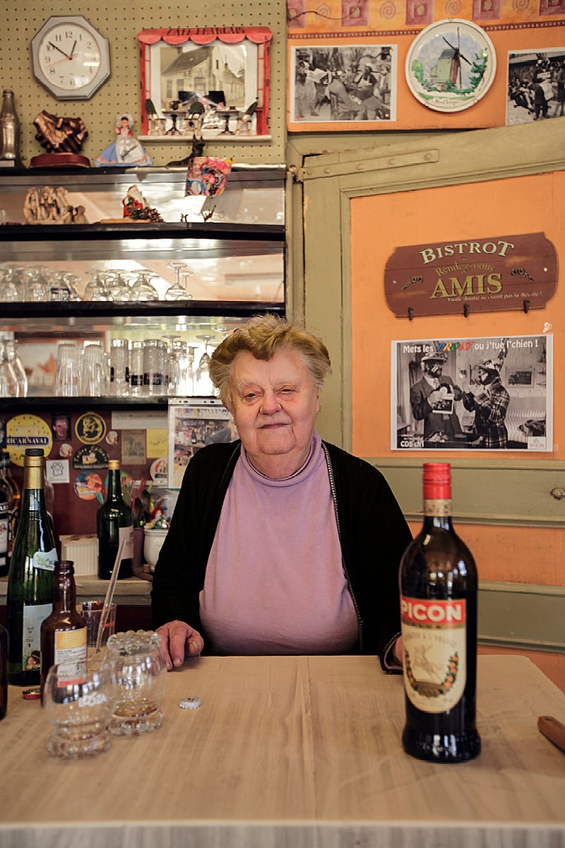 Elderly woman seated behind a wooden table inside the pub, with bottles, glasses, old signs, and a bottle of Picon in the foreground.