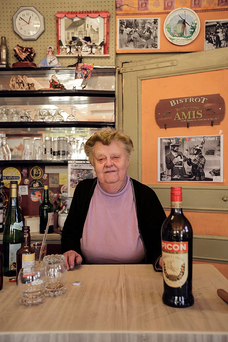 Elderly woman seated behind a wooden table inside the pub, with bottles, glasses, old signs, and a bottle of Picon in the foreground.