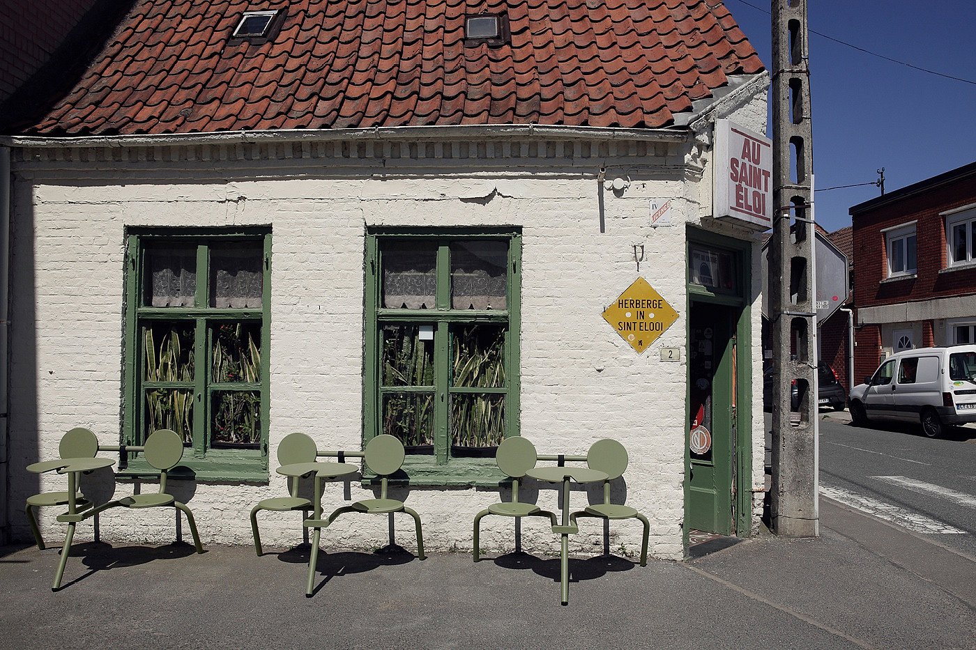 Drie saliegroene Bistroo tweezitters stonden opgesteld tegen de witte bakstenen gevel van Au Saint-Eloi, met groen geschilderde ramen, ingegoten sansevieria's achter het glas en het pubbord boven de hoekingang.