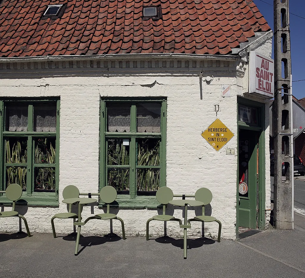 Trois bistroos vert sauge à deux places alignés contre la façade en briques blanches d’Au Saint-Eloi, avec des fenêtres peintes en vert, des sansevierias en pot derrière le verre et le panneau du pub au-dessus de l’entrée d’angle.