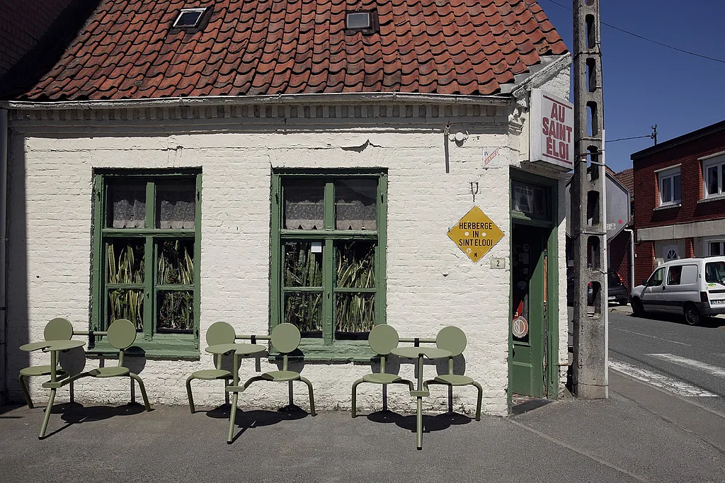 Drie saliegroene Bistroo tweezitters stonden opgesteld tegen de witte bakstenen gevel van Au Saint-Eloi, met groen geschilderde ramen, ingegoten sansevieria's achter het glas en het pubbord boven de hoekingang.