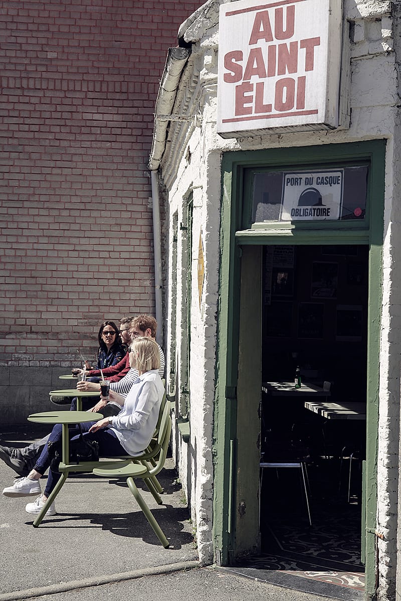 Several people sitting on Bistroo seats outside the pub in a neat row, enjoying drinks along the sunlit white façade beside the open doorway.