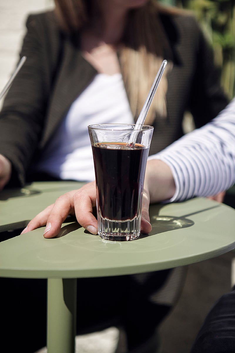Close-up of a picon in a tall glass resting on the rounded tabletop of a sage-green Bistroo seat, with two people softly blurred in the background.