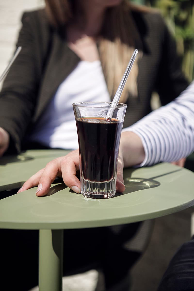 Close-up of a picon in a tall glass resting on the rounded tabletop of a sage-green Bistroo seat, with two people softly blurred in the background.
