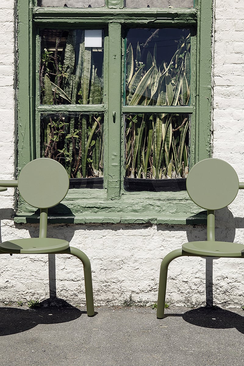 Two empty sage-green Bistroo chairs placed beneath a green-painted window filled with tall sansevieria plants inside the pub.