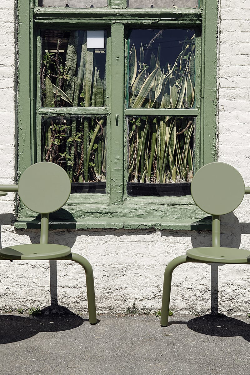 Two empty sage-green Bistroo chairs placed beneath a green-painted window filled with tall sansevieria plants inside the pub.
