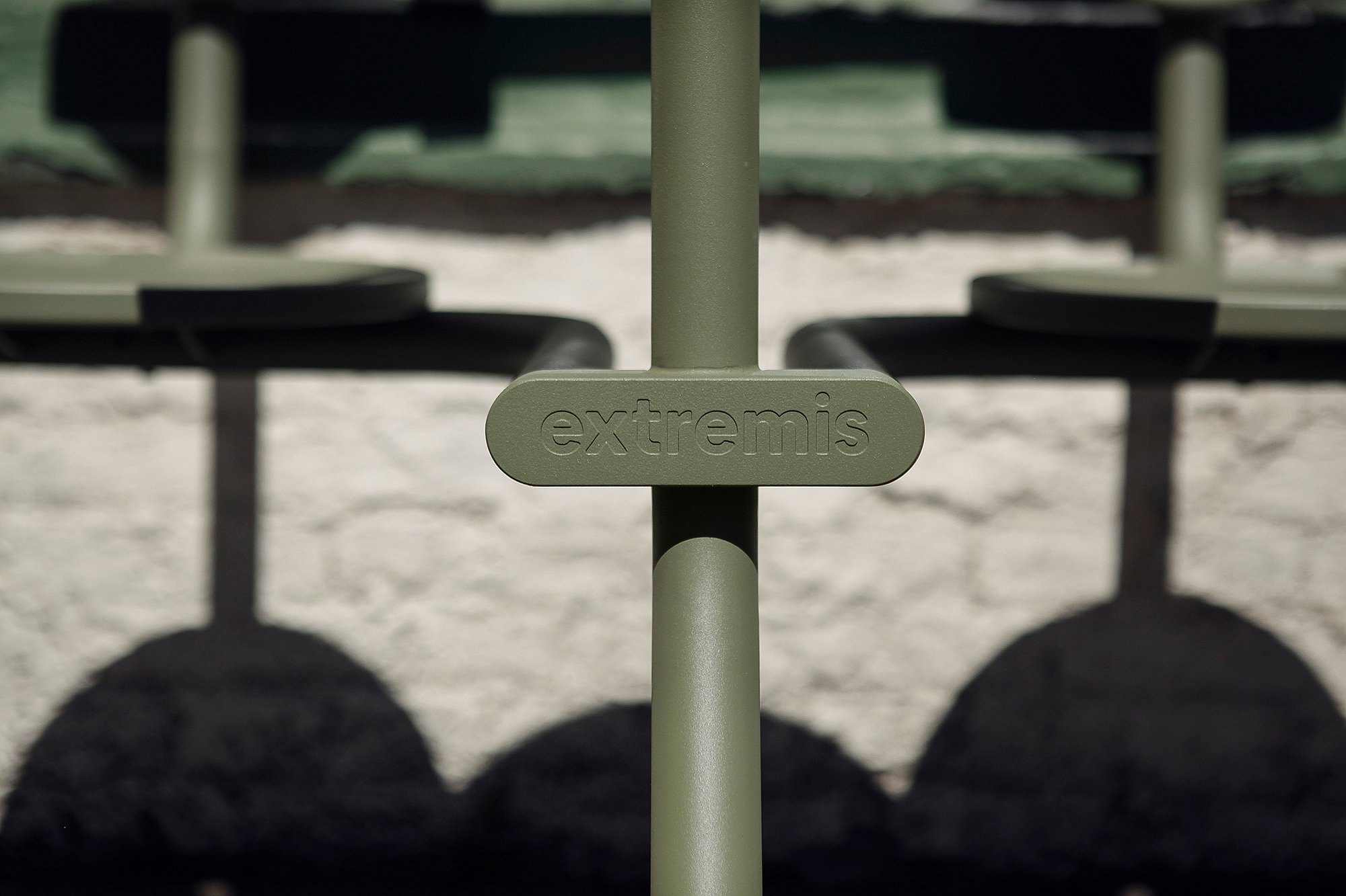 Close-up of the embossed Extremis logo on the vertical support of a sage-green Bistroo seat, with the round seats and their shadows blurred behind