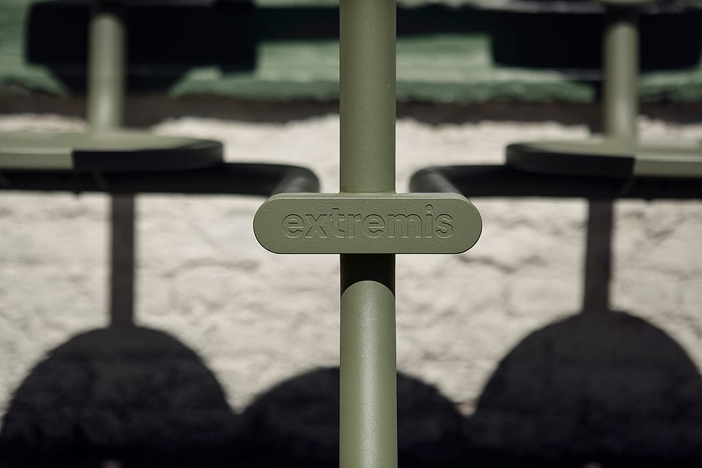 Close-up of the embossed Extremis logo on the vertical support of a sage-green Bistroo seat, with the round seats and their shadows blurred behind