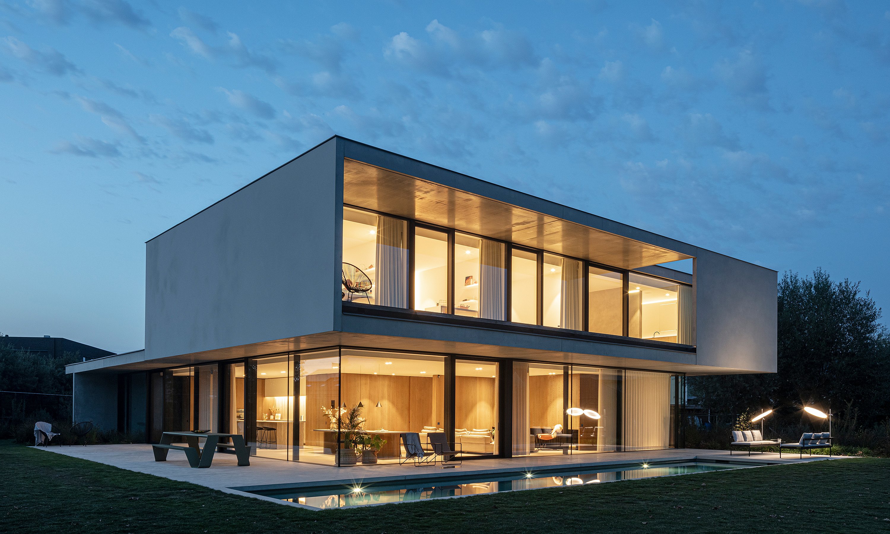 The modern villa glowing at dusk with the pool in front, a Hopper picnic table on the terrace, and Sol+Luna loungers lit beside the garden.