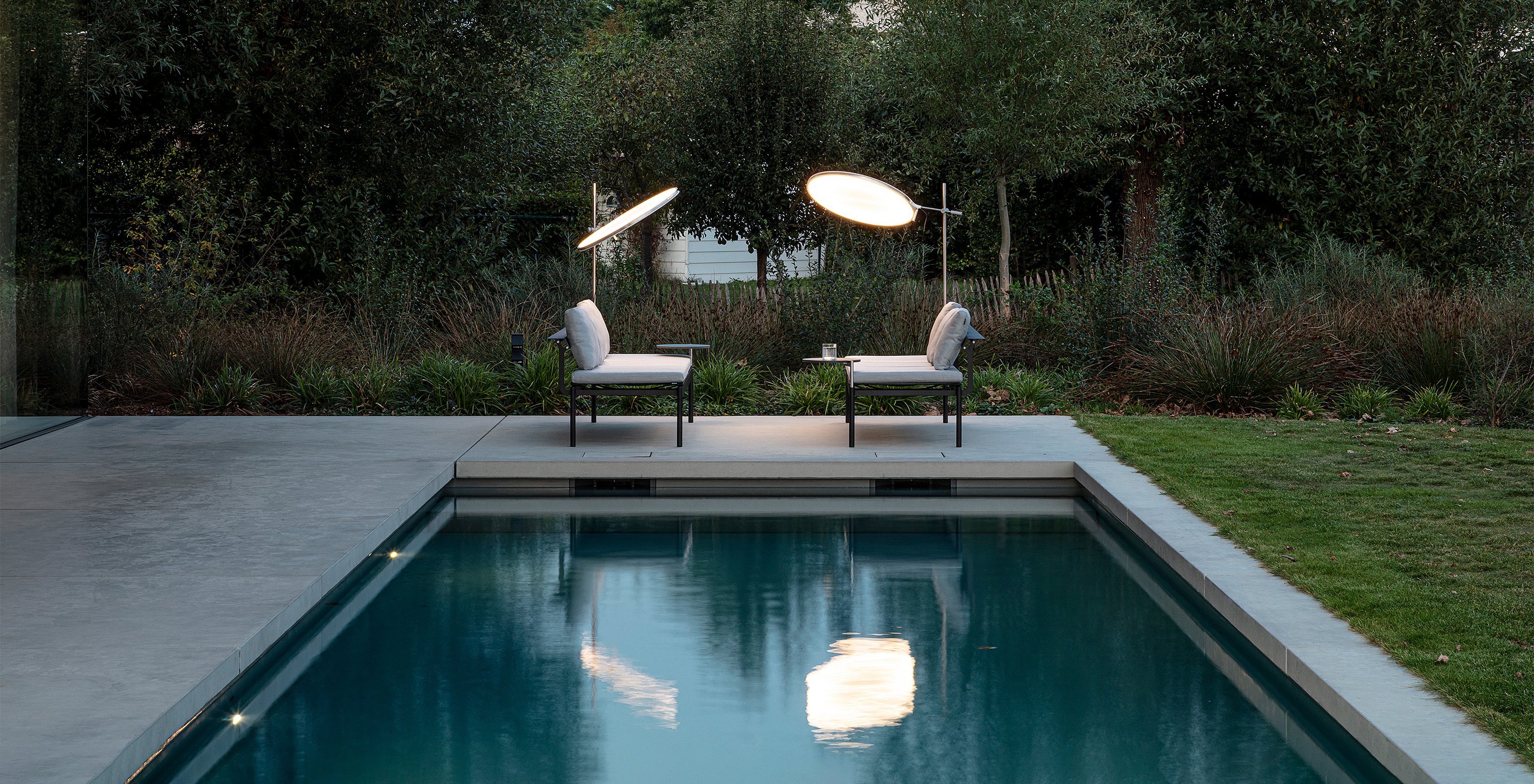 Two illuminated Sol+Luna loungers facing each other across the far end of the pool at dusk, their glowing shades reflected in the water.