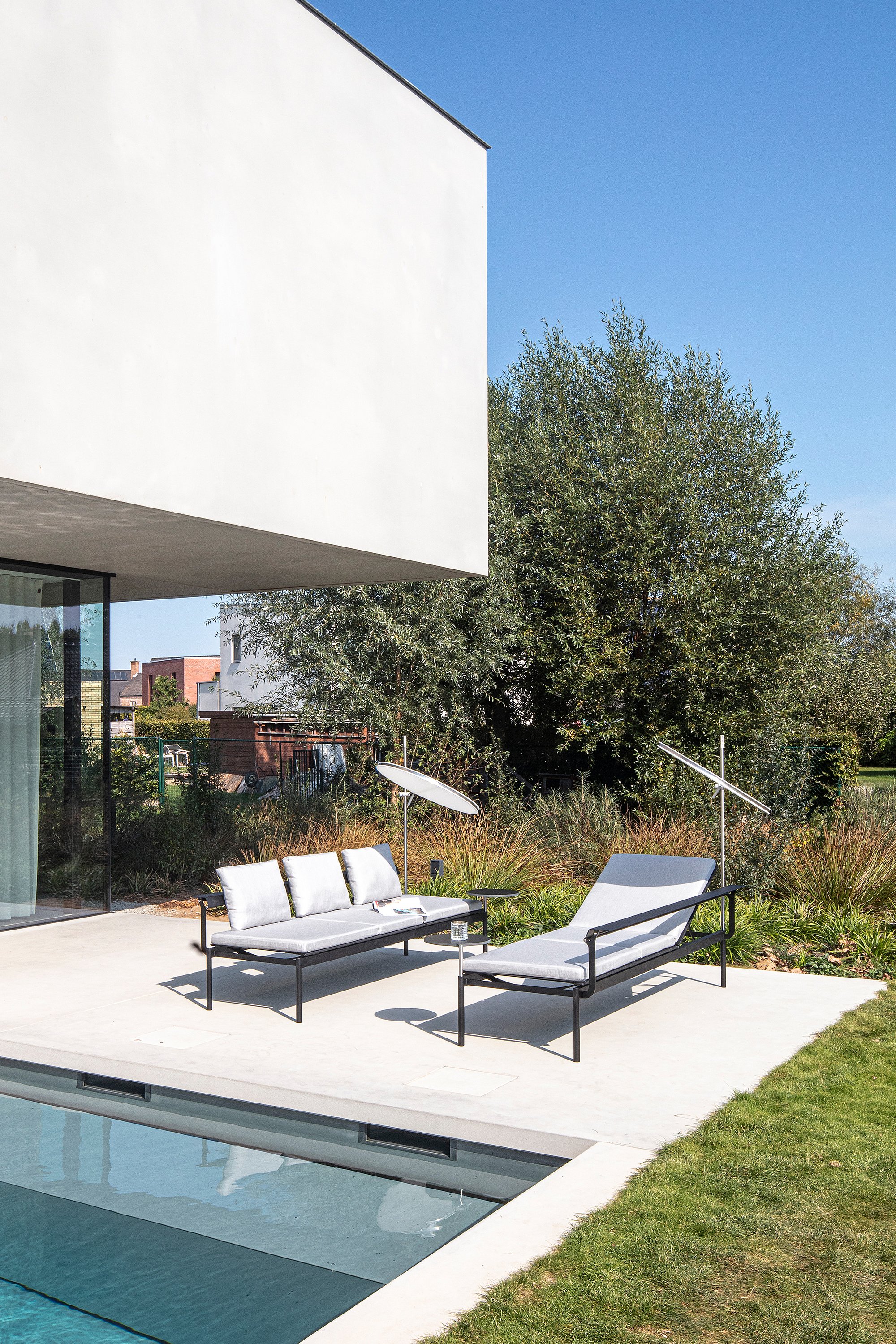 Two Sol+Luna loungers with light cushions and oval shades arranged on the pool terrace beside the villa’s glass façade and planted garden border.