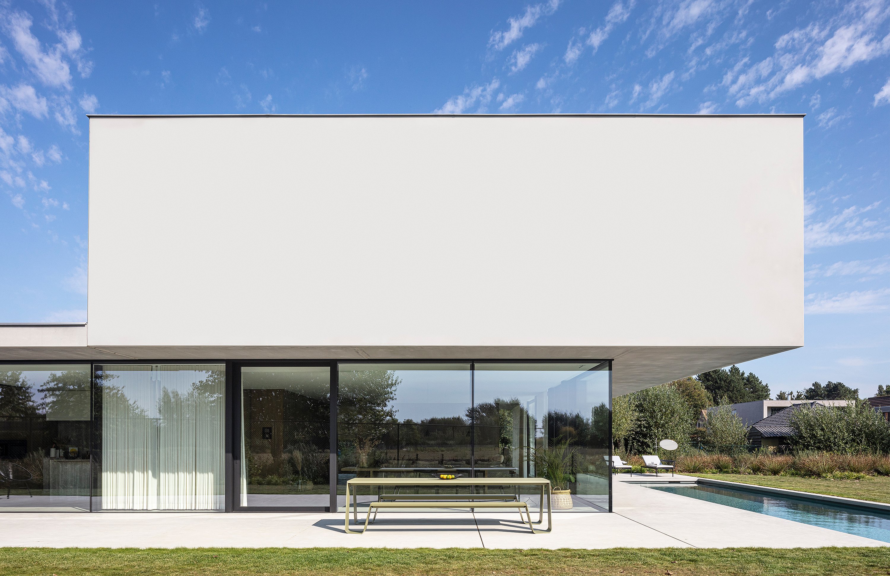 Reed-green Hopper picnic table placed on a concrete terrace beside floor-to-ceiling windows and a long swimming pool, set against the clean lines of a minimalist white villa.