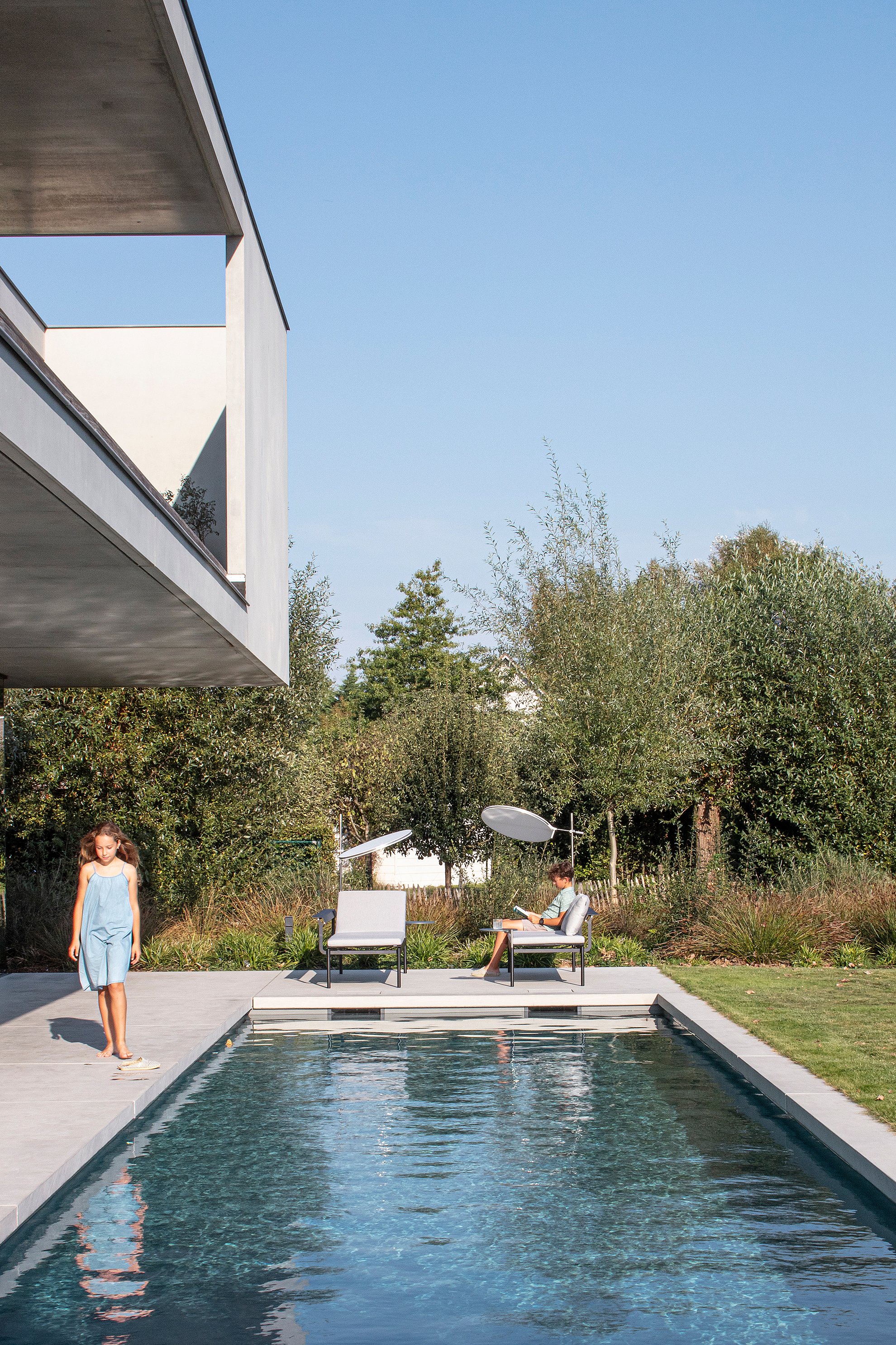 “Long rectangular swimming pool beside the house, with a girl walking along the edge and a boy relaxing near two Sol+Luna loungers in the garden.