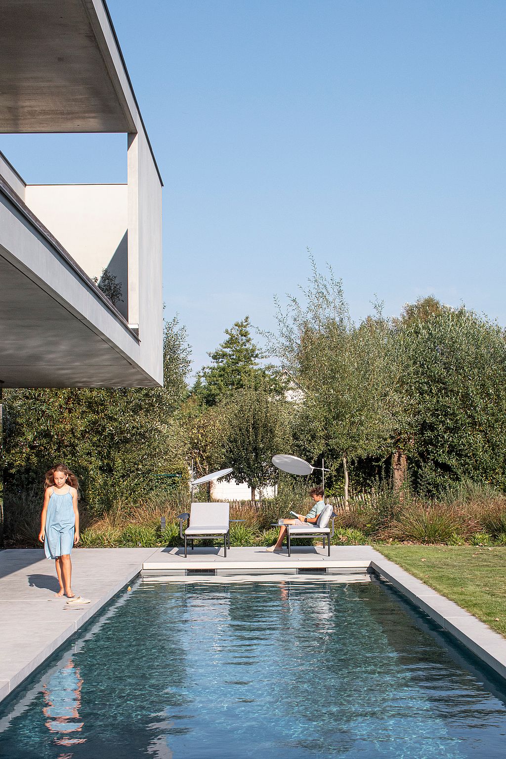“Long rectangular swimming pool beside the house, with a girl walking along the edge and a boy relaxing near two Sol+Luna loungers in the garden.
