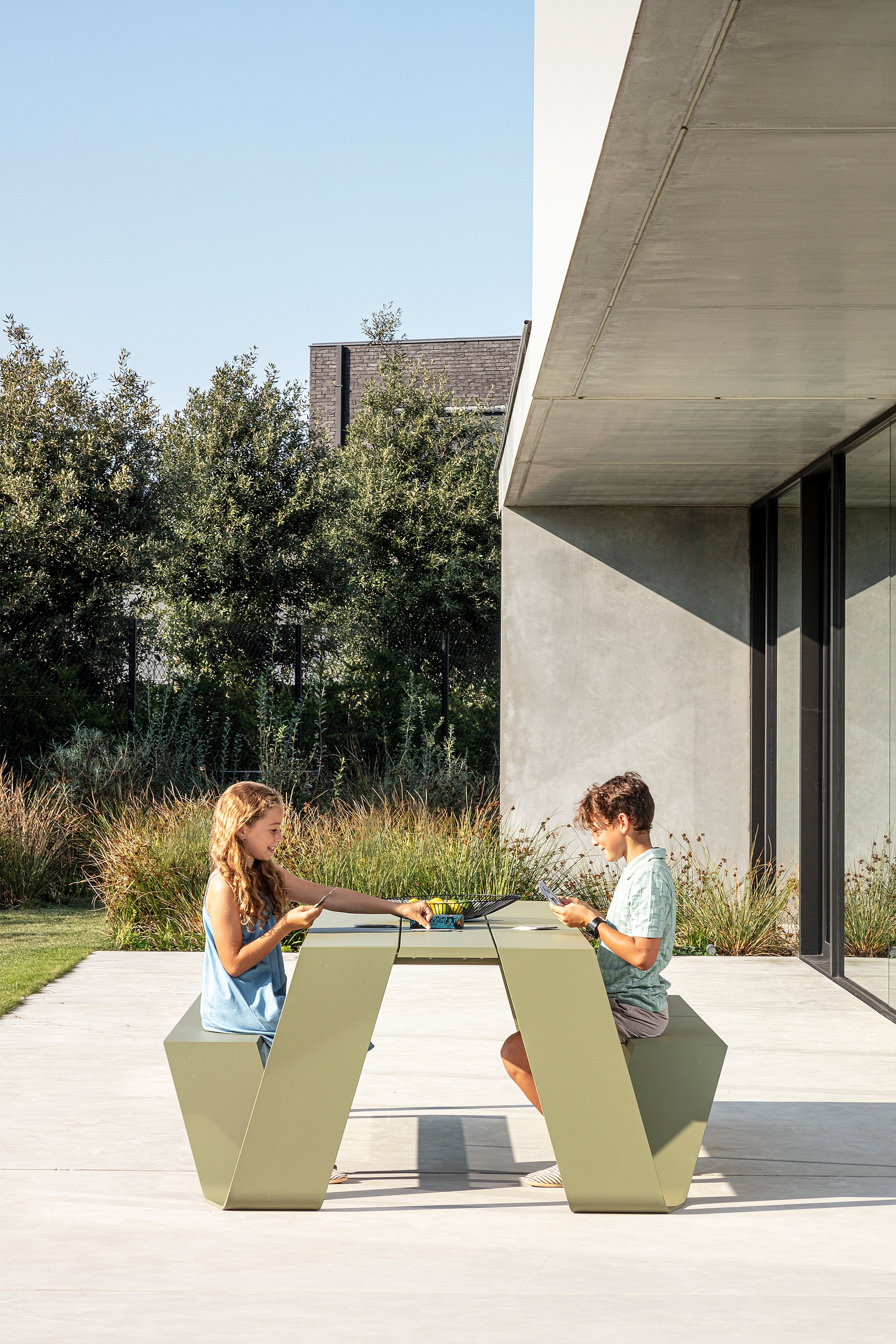 Two children sit opposite each other at a reed-green Hopper picnic table on the sunny terrace, playing cards beside the modern house.