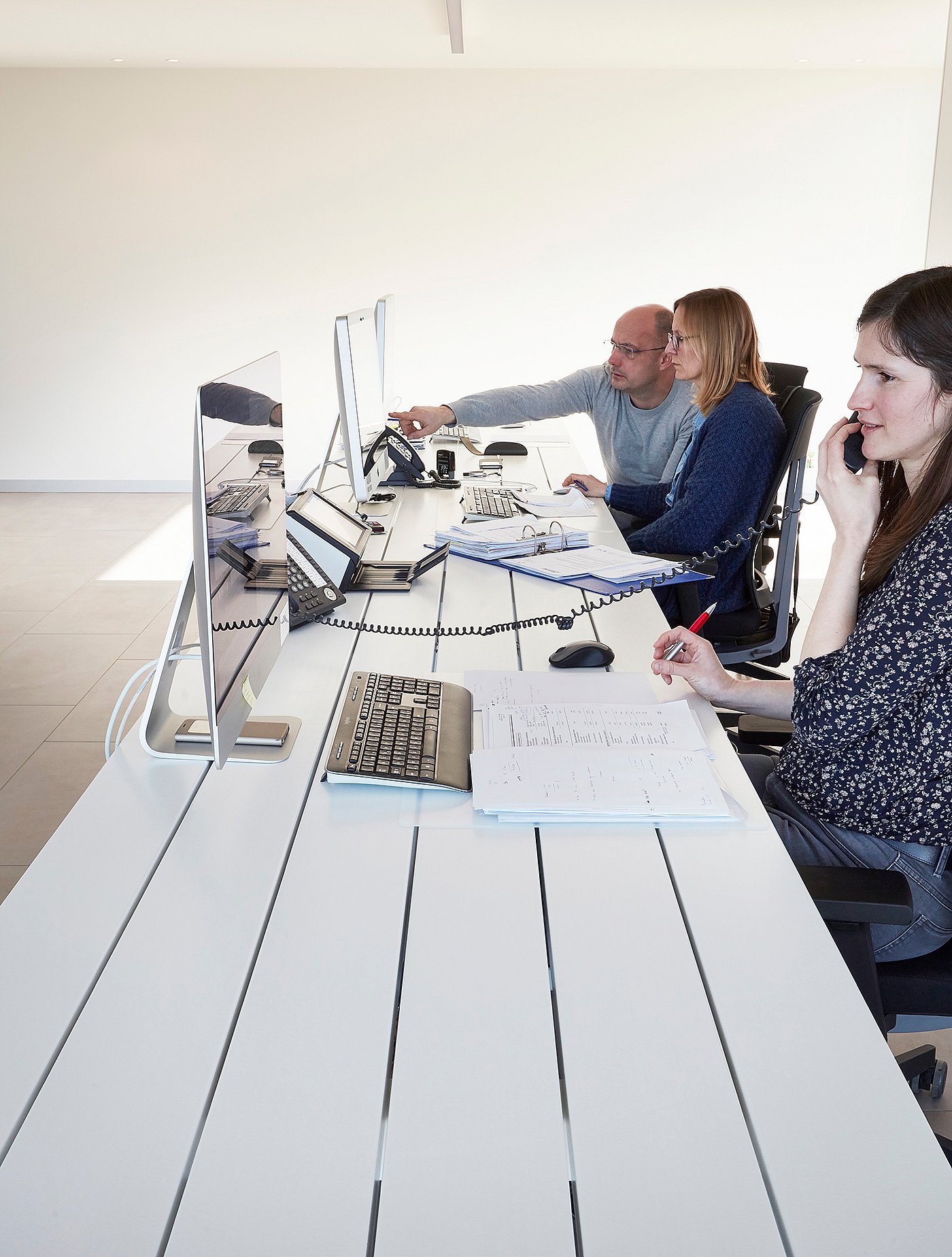 Drei Mitarbeiter arbeiten Seite an Seite an einem langen weißen Marina-Schreibtisch, Computer, Papierkram und Telefone sind auf der Tischplatte angeordnet.
