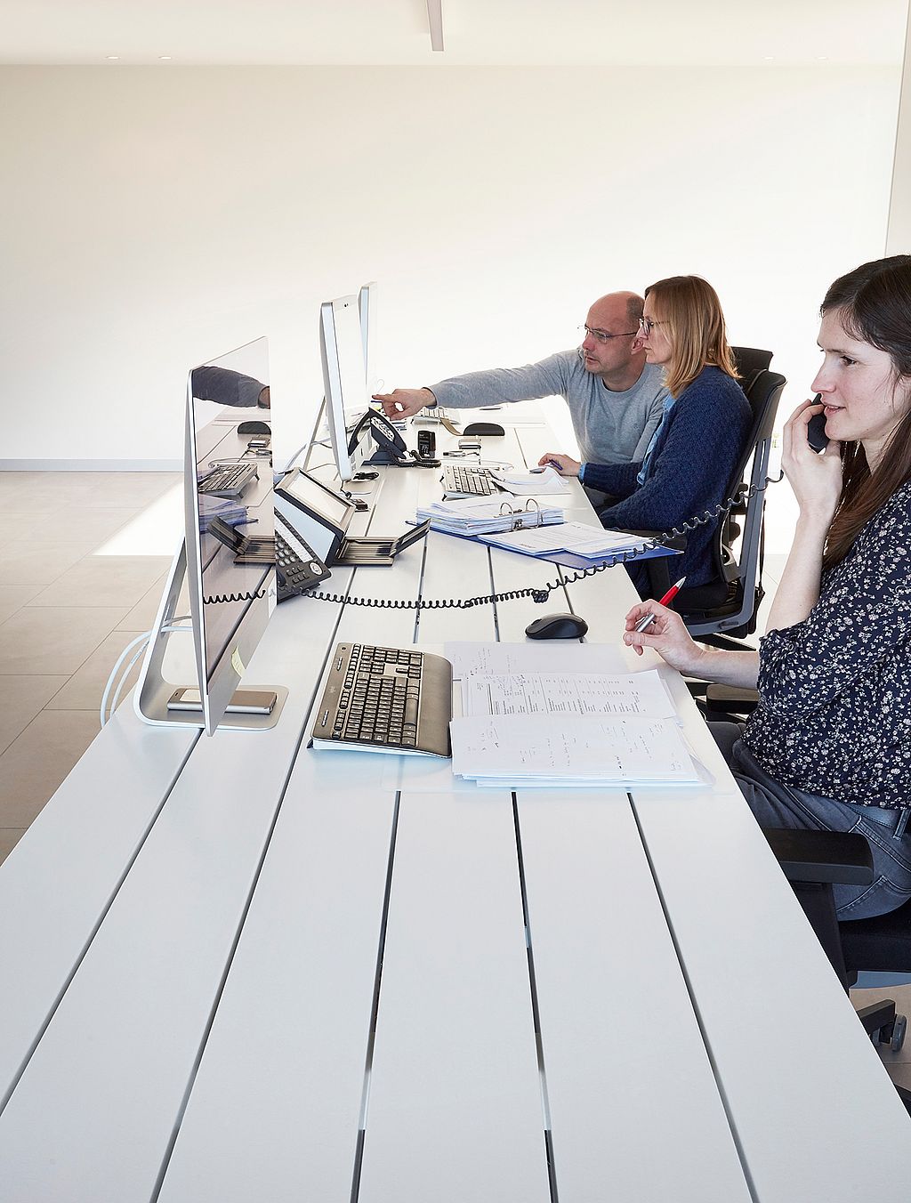 Drei Mitarbeiter arbeiten Seite an Seite an einem langen weißen Marina-Schreibtisch, Computer, Papierkram und Telefone sind auf der Tischplatte angeordnet.