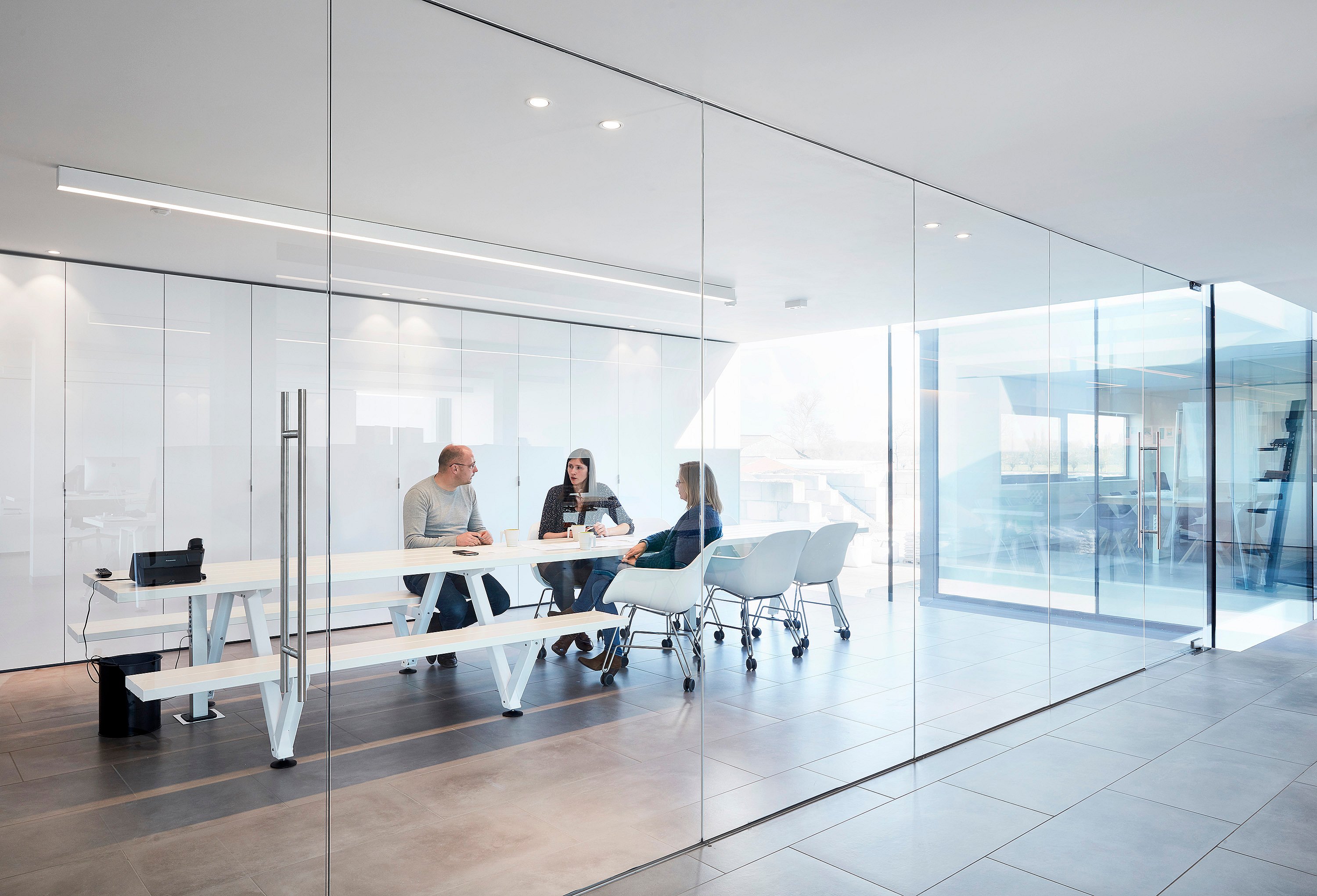 Minimal glass-walled meeting room with a long white Marina Combo table and matching bench, where three colleagues sit together on white Captain’s Chairs in a bright modern office.