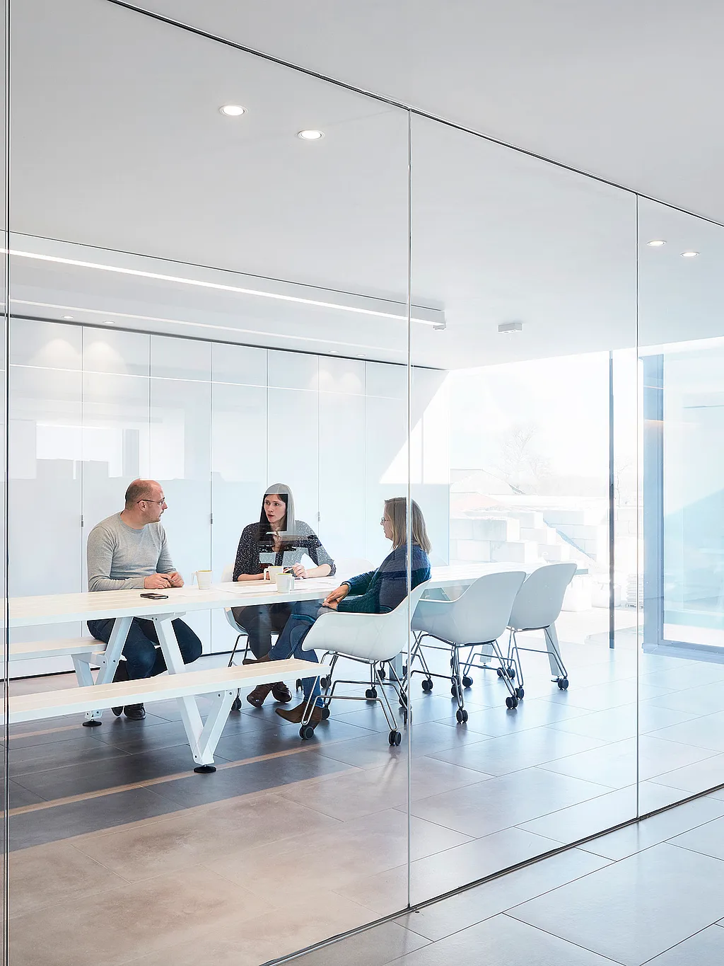 Minimal glass-walled meeting room with a long white Marina Combo table and matching bench, where three colleagues sit together on white Captain’s Chairs in a bright modern office.